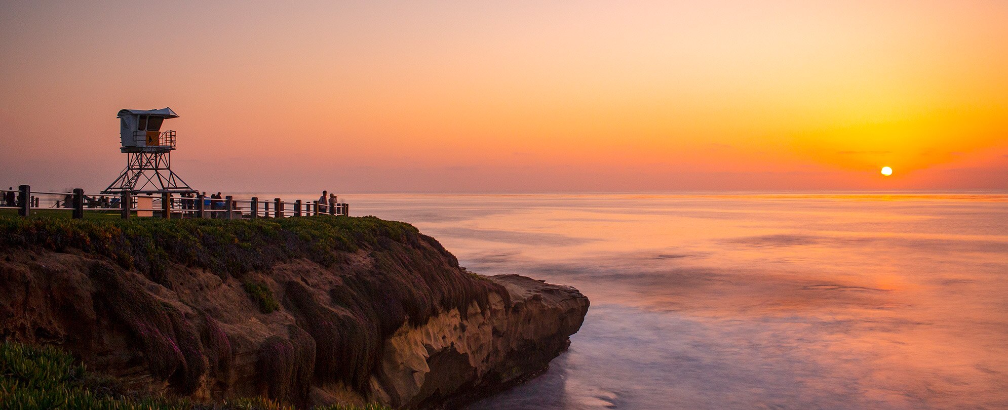 Silhouetted spectators watch the sun set over the ocean and rocky cliff.