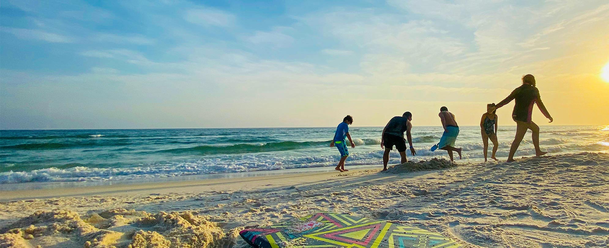 A family of five walks around the beach playing in the sand along the shore with a colorful boogie board in the foreground.