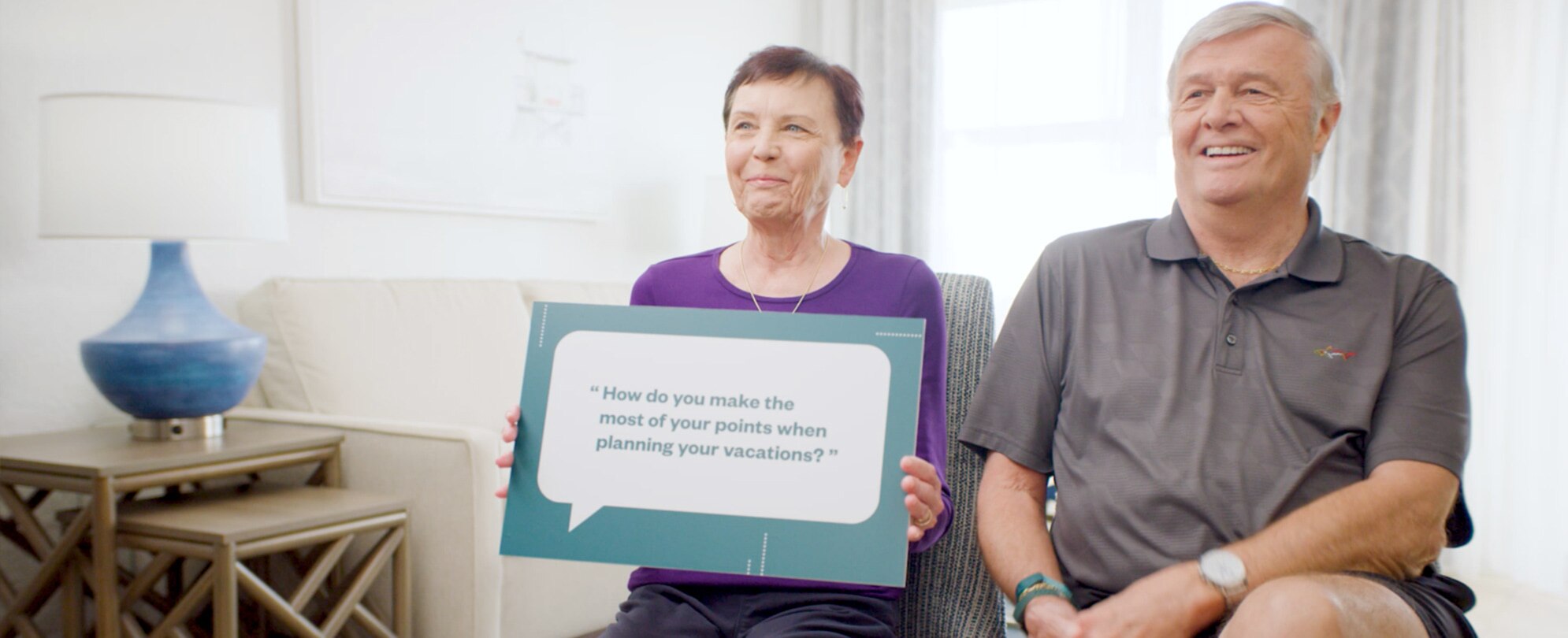 An older woman sitting next to her husband, she is holding a sign that says “How do you make the most of your points when planning your vacations?”