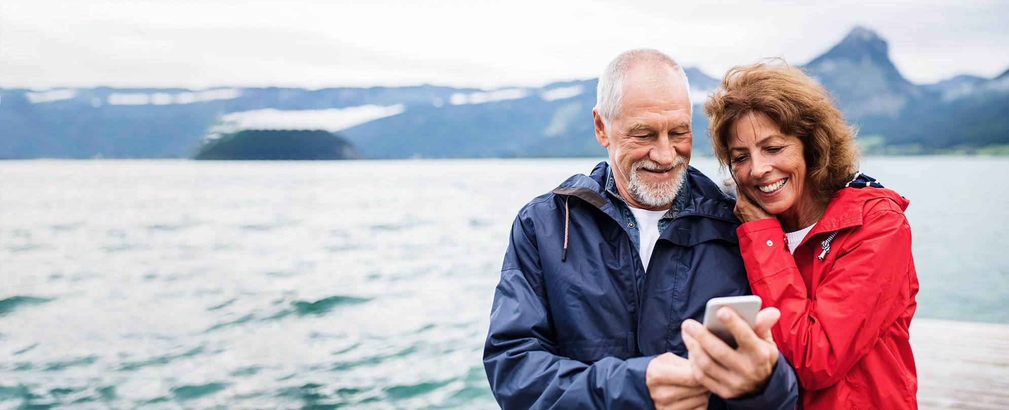 A woman with her arms on her husband's shoulder as he takes a photo of them with the snow-covered mountains in the background.